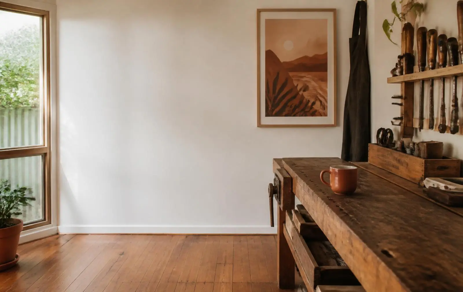 A calm, sunlit small-workshop interior with a warm timber workbench, off-white walls, and a single terracotta mug.